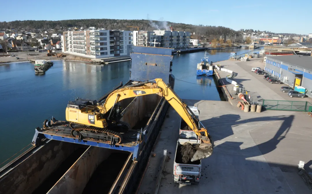 Gravemaskin plassert på bru over båt losser de første massene på kaia i Tønsberg en tidlig morgen i soloppgang