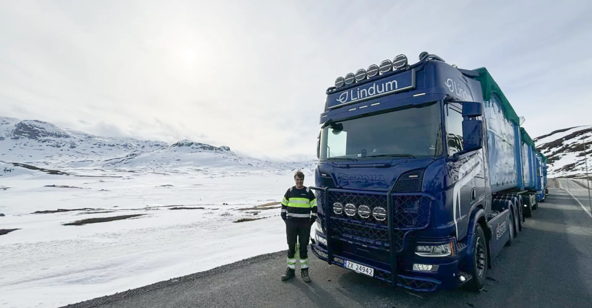 Mann poserer foran vogntog på vei med snølagde fjell på begge sider.